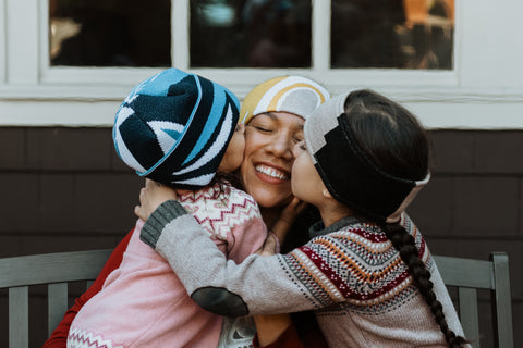 Two young Native children, a daughter wearing a wool hat and a son wearing a wool headband, hug their mom and kiss her cheek.
