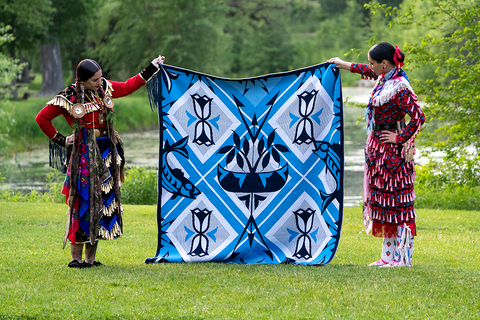 Two women in beautiful Ojibwe regalia hold a blue and white wool blanket in a green meadow