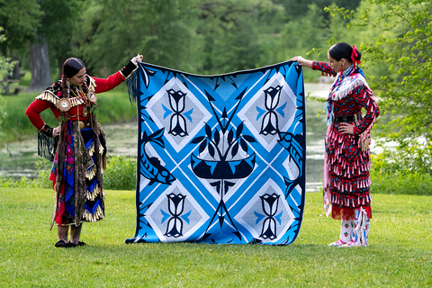 Two women in jingle dress regalia stand in a green field holding a blue and white wool blanket.