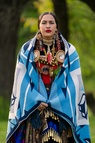A woman in regalia stands facing the camera. Her black hair is in braids, and she is wrapped in a blue and white Good Life Blanket