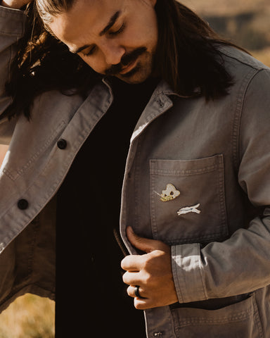 A man with dark hair and a dark mustache wears a jacket that has a wolf and buffalo enamel pin on it.