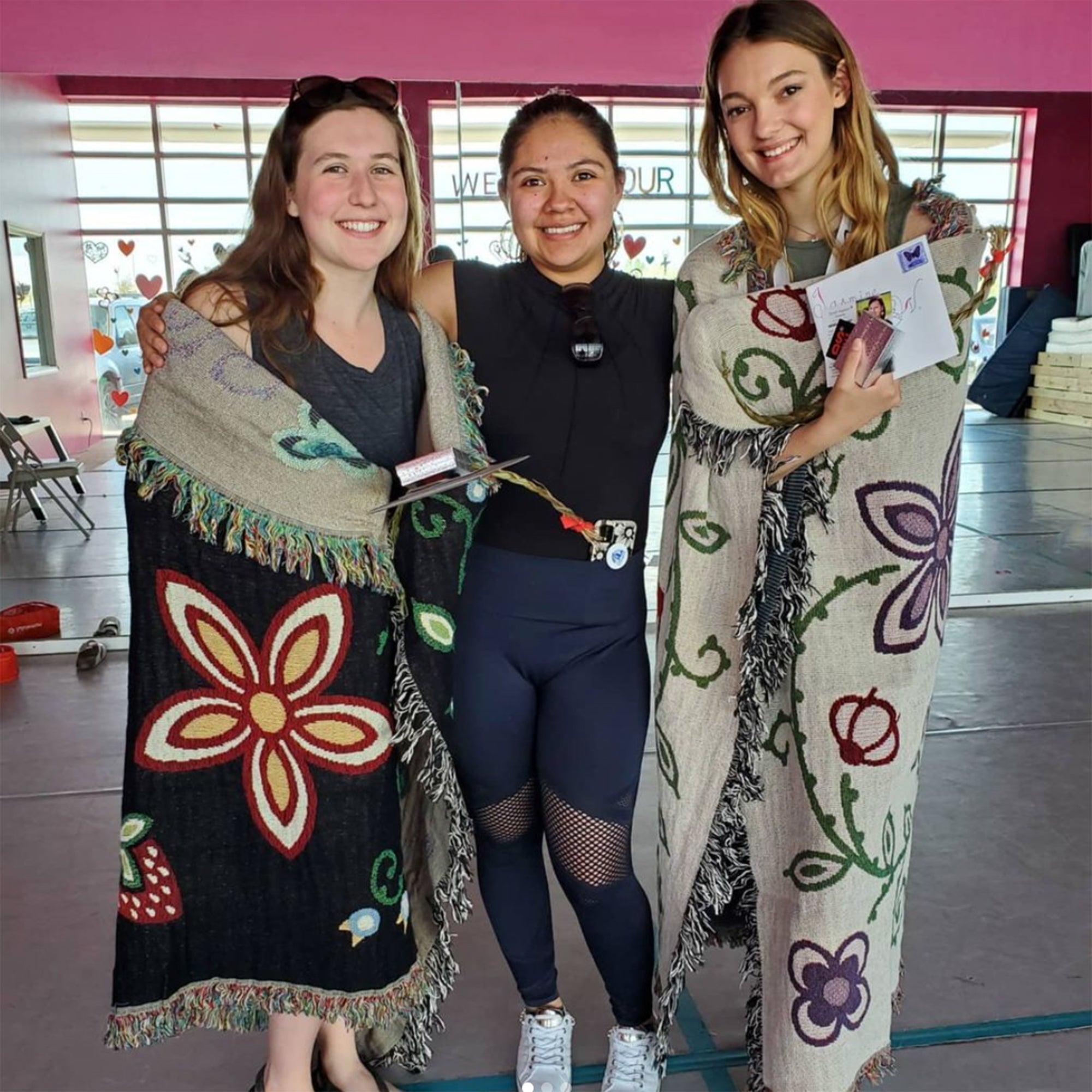 Three-women-stand-smiling-and-graduates-on-either-end-are-wrapped-in-a-black-and-a-beige-throw-blanket-with-woodland-floral-designs