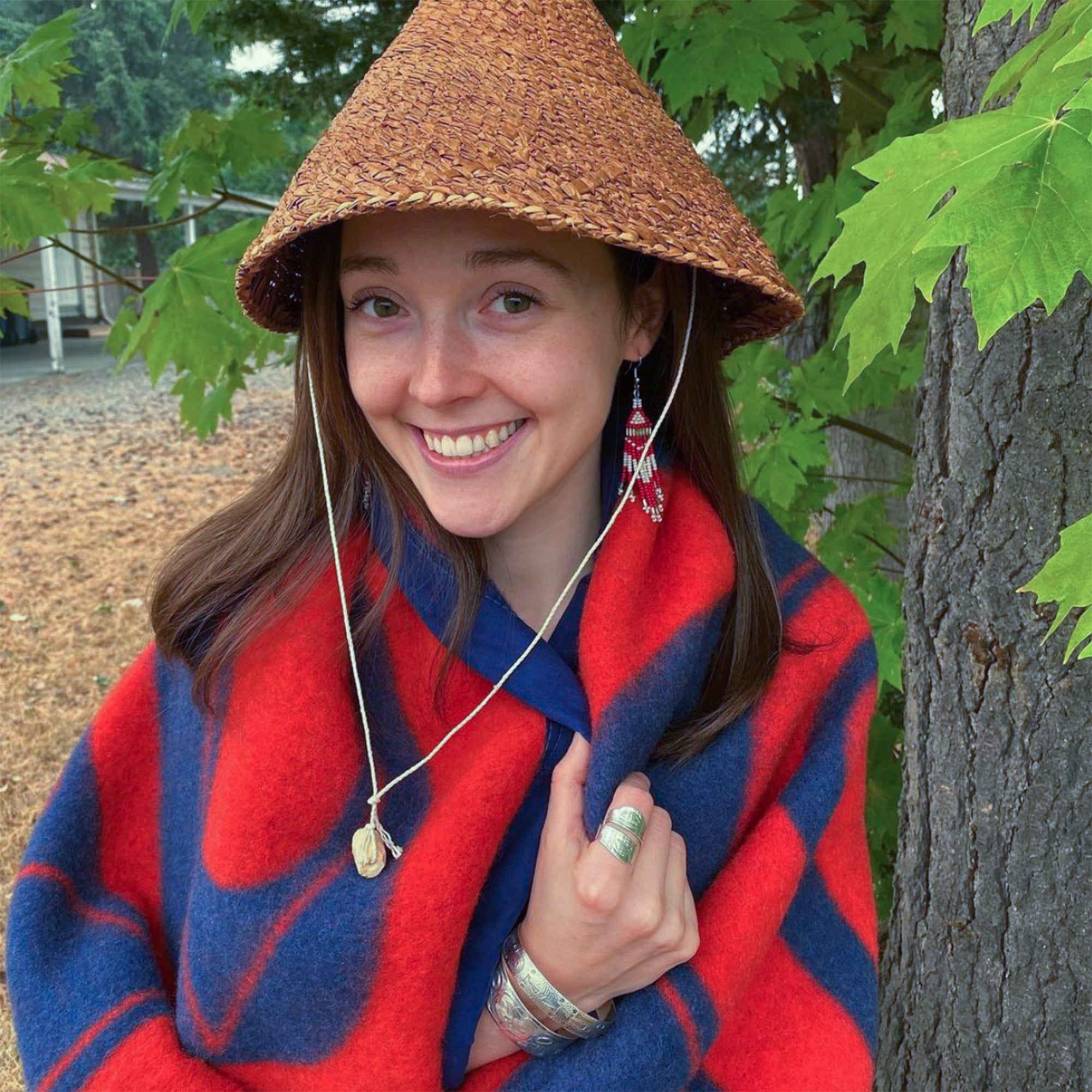 A-woman-with-a-cedar-hat-smiles-and-stands-wrapped-in-a-red-and-blue-wool-blanket-with-Northwest-Coast-designs.