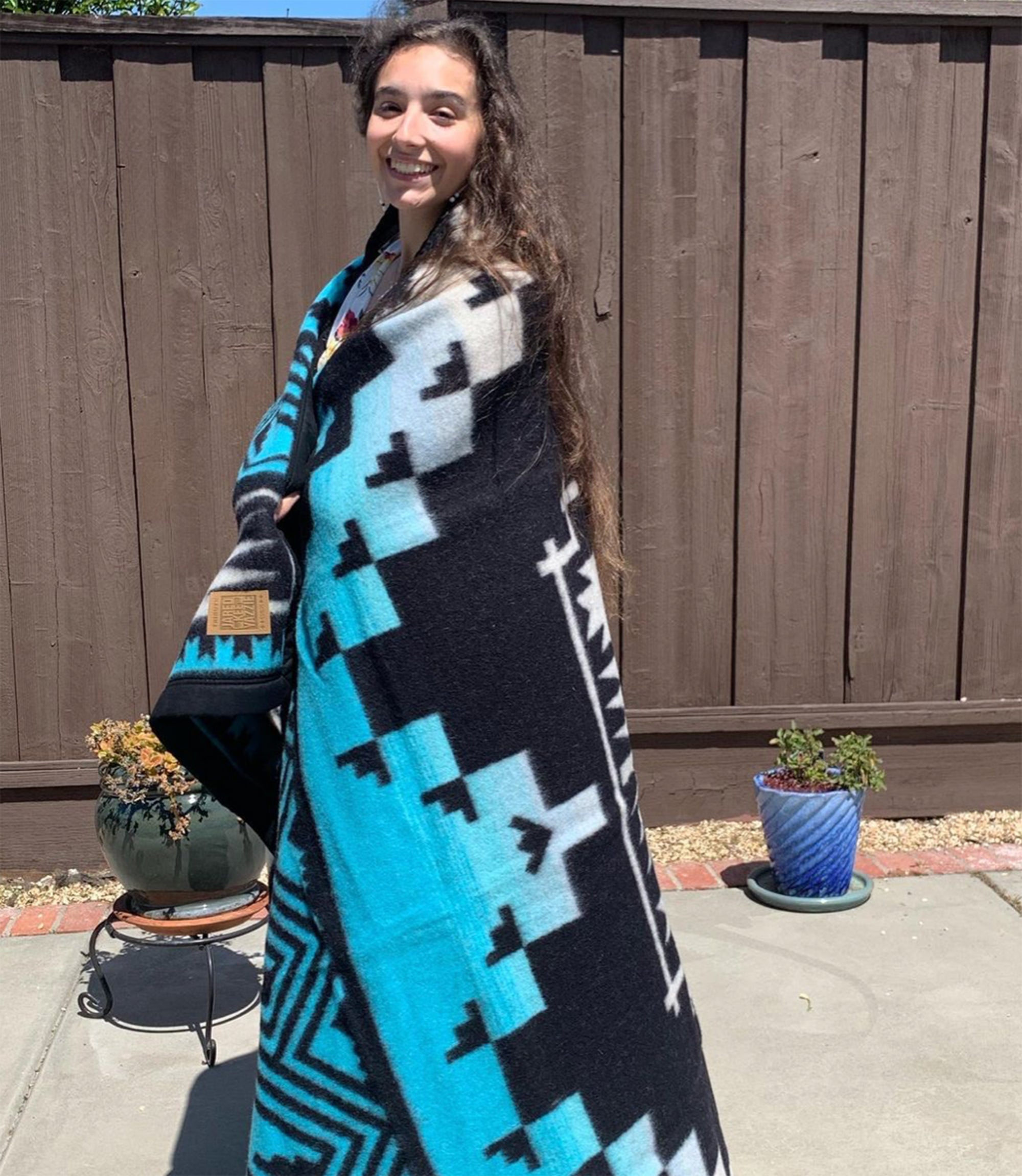 A-woman-with-long-brown-hair-stands-smiling-and-wrapped-in-black-and-turquoise-wool-blanket