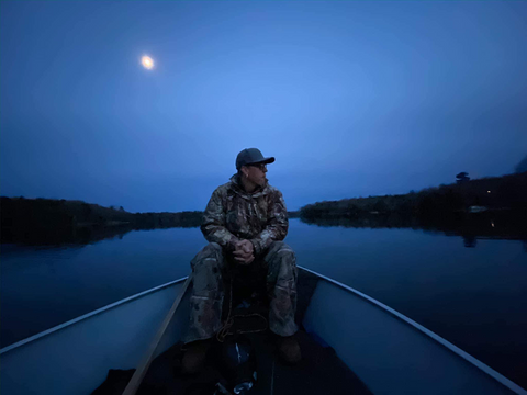 A man sits on the prow of a canoe at dusk. A full moon is seen over his shoulder on the left side of the image, while black hills rise around him.