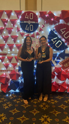Two smiling Native American women in black dresses holding awards in front of a wall with "40 Under 40" banners.