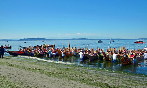 A long row of canoes lines up on a Puget Sound shoreline during Stephanie Masterman (Tlingit) sits in the center of the canoe waring a purple life jacket and holding her paddle during the Power Paddle to Puyallup 2018. Photo courtesy of Chris Stearns (Navajo)