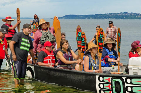 A Northwest Coast tribal canoe sits in the water on a Pacific beach in 2018. The canoe is full of Native American, Alaska Native, and First Nations pullers, who sit with their paddles.
