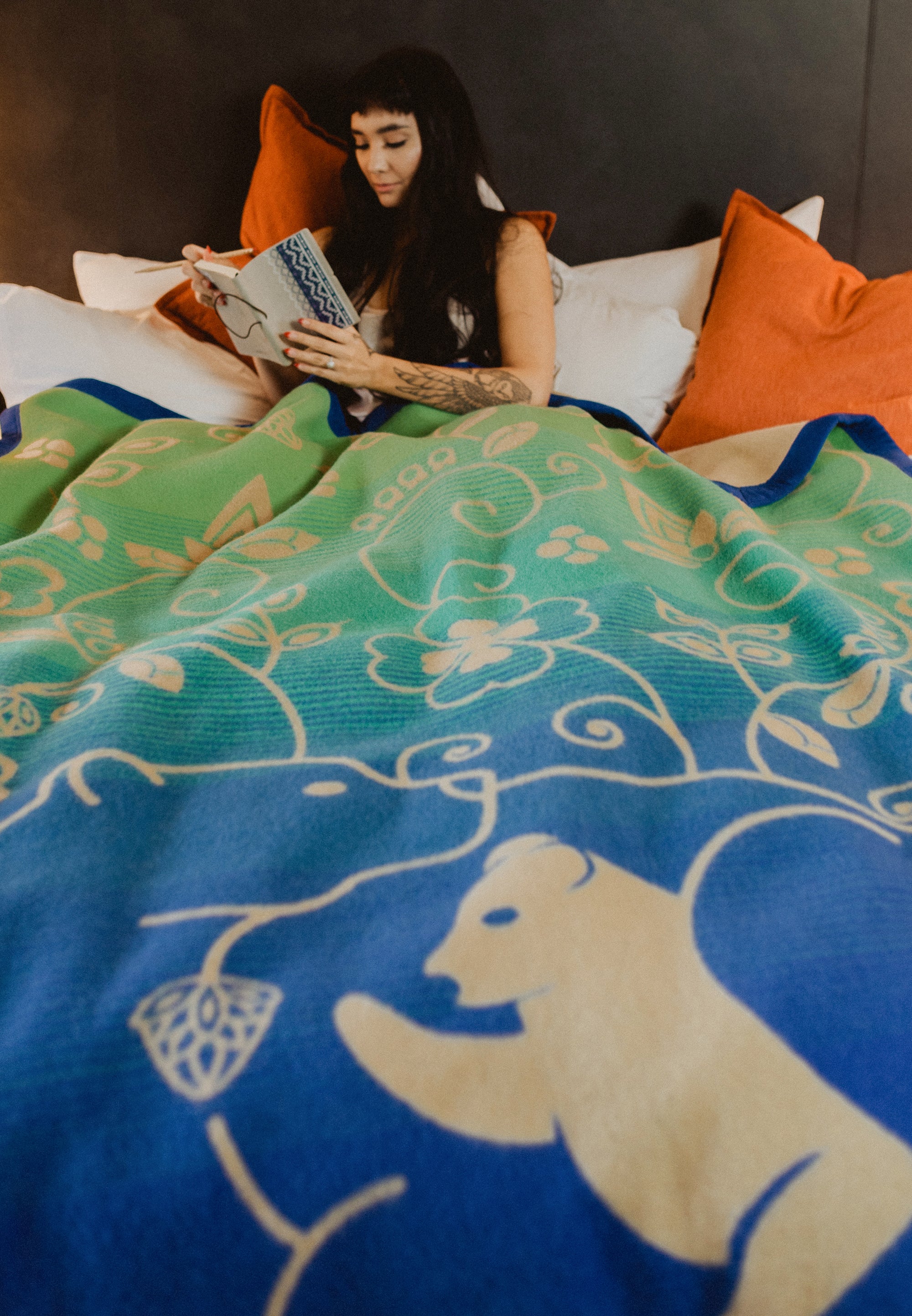 Woman-reads-in-bed-with-blue-tan-and-green-wool-blanket-covering-her.-Blanket-has-bear-and-cub-design-surrounded-by-woodlands-floral