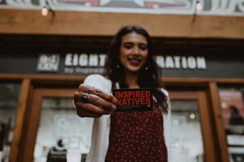 A woman holds a small sign toward the camera, which reads "I support Inspired Natives, not "Native-Inspired."