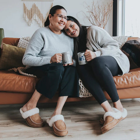 Two Indigenous women sit on a couch wearing Manitobah slippers