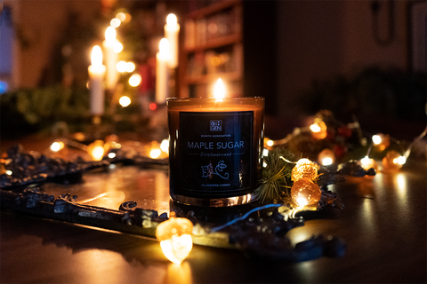 A Maple Sugar Candle in a brown glass jar sits lit on a dark wood table with other lit candles in the background