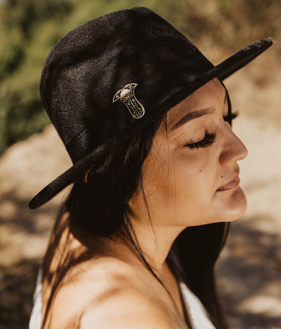 A photo of a woman in profile. She wears a black hat and her long dark hair streams over her shoulder. 