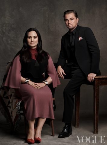 A Native American woman in a mauve dress sits next to a man wearing a black suit, also seated