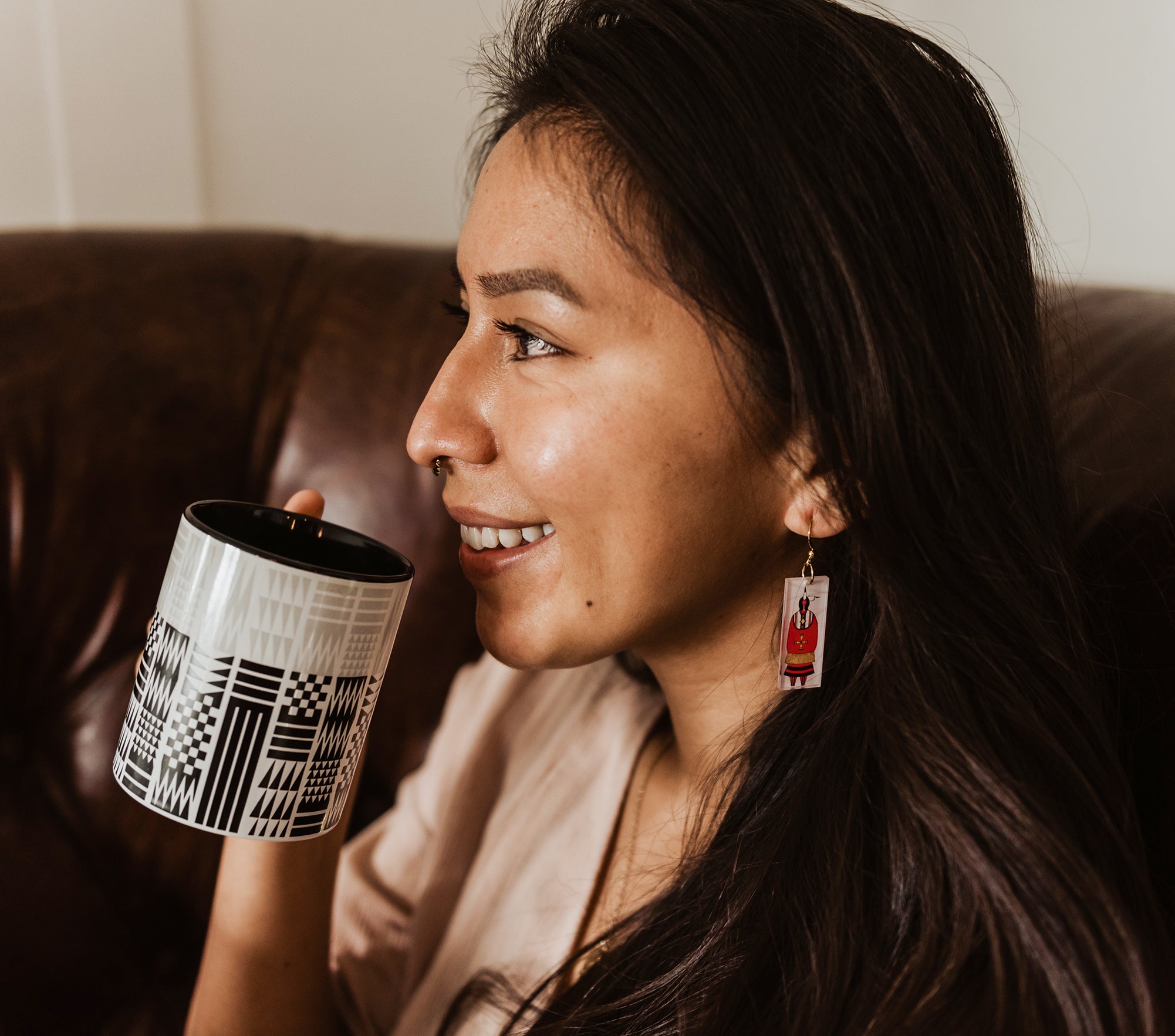 Woman-smiles-holding-black-and-white-Coast-Salish-Pattern-mug-and-wears-white-earrings-with-woman-in-red