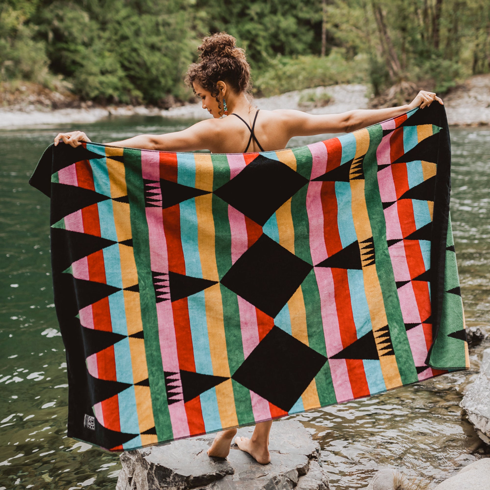 Woman-stands-on-rock-near-water-and-holds-towel-out-behind-her-to-display-the-green-pink-red-blue-orange-ribbons-design