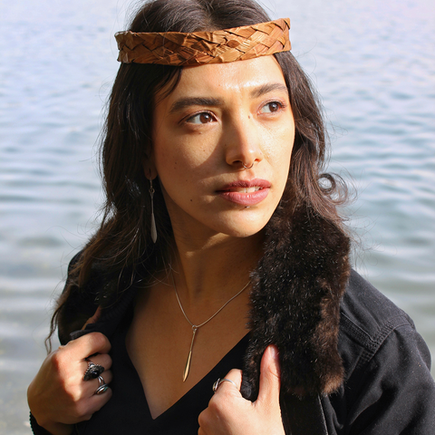 A Tlingit woman with a cedar headband and septum piercing models earrings and a necklace in the shape of small silver paddles