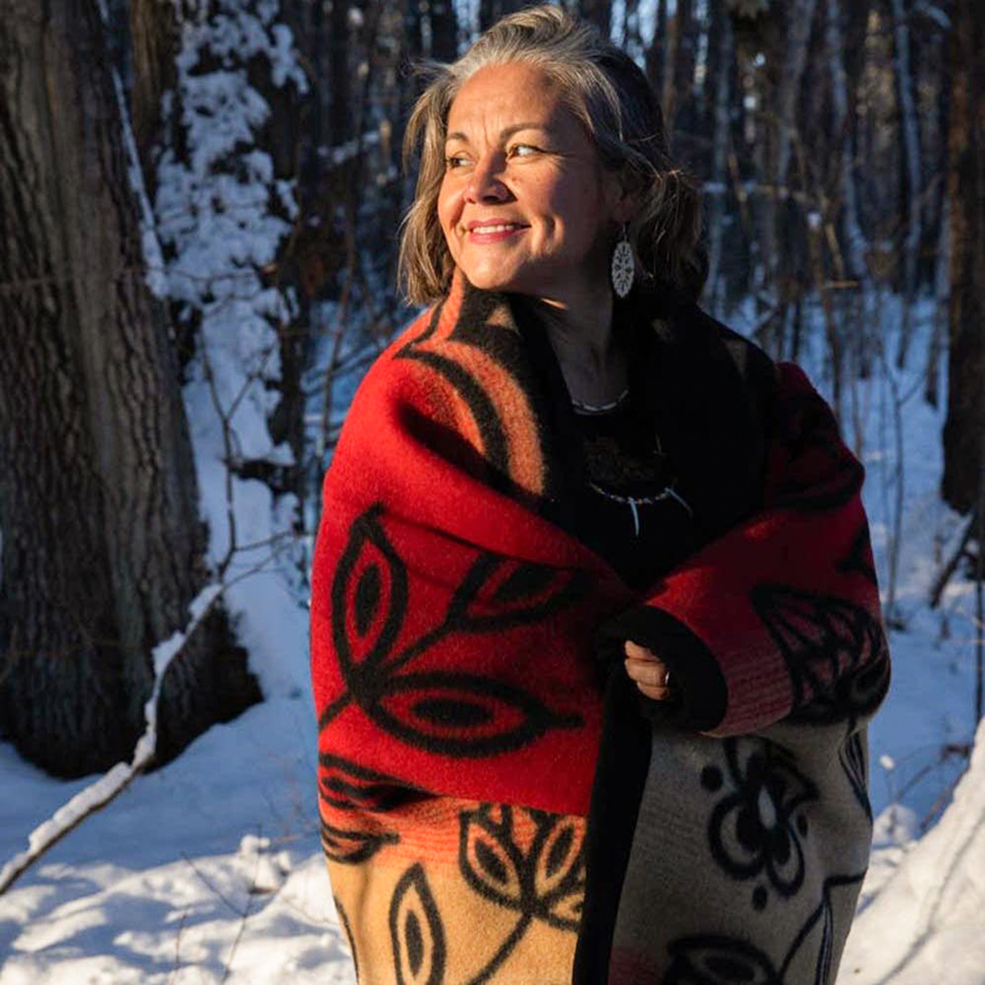 Anishinaabe-arts-entrepreneur-Sarah-Agaton-Howes-stands-in-snow-wrapped-in-black-and-red-renewal-wool-blanket-and-oval-floral-birch-bloom-earrings