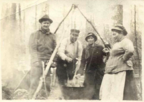 A historic photo of four Anishinaabe people standing around a pole-frame cooking lift and a pot of wild rice