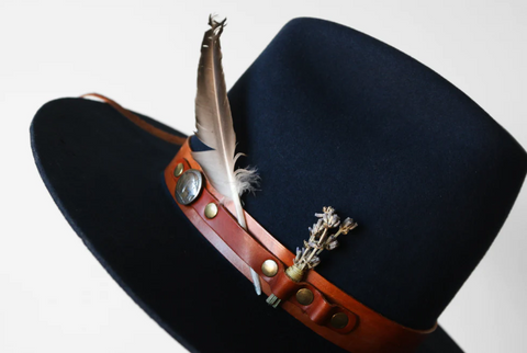 A black wide-brimmed hat has a brown leather hat band and feather