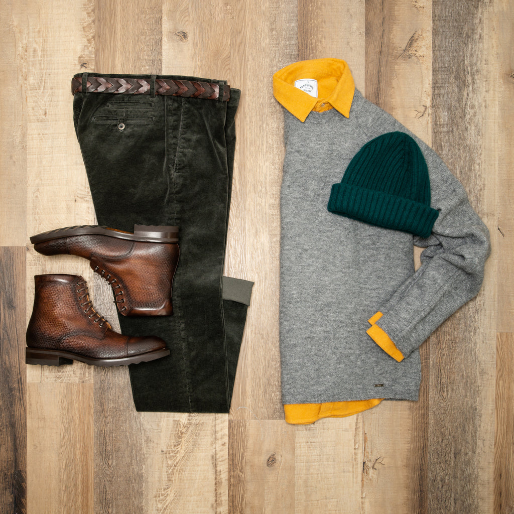 Flat lay image of olive corduroy trousers, brown boots, grey sweater with mustard portuguese flannel shirt under, and a green toque