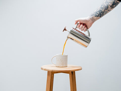 A tattoed hand pouring French press coffee into a mug on a stool.jpg