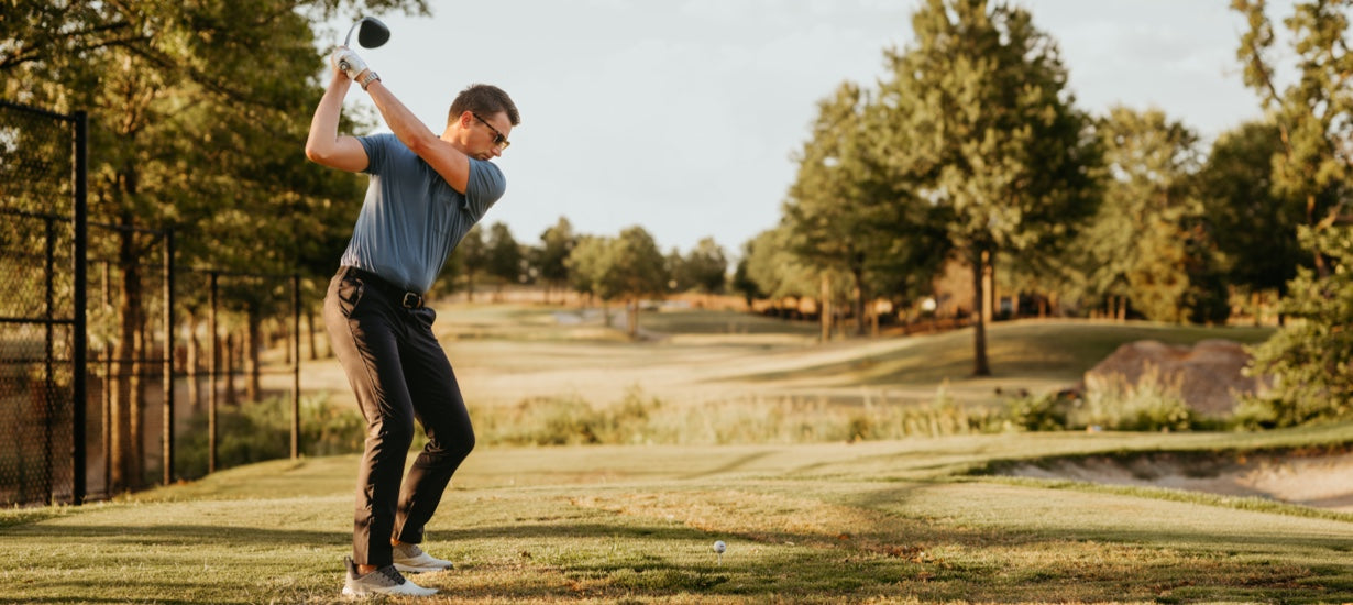 A young man swinging a golf club while wearing performance pants and a technical polo.