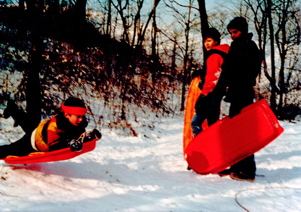 A vintage photo of three young boys wearing orange + black while sledding in the snow.