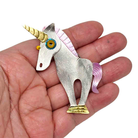 Unicorn brooch created by Chickenscratch Jewelry, shining majestically with a silver-plated brass body, gold leaf hooves and horn, pinkish purplish mica power-coated mane and tail, and a sparkly blue eye disc around a cast resin yellow eye.