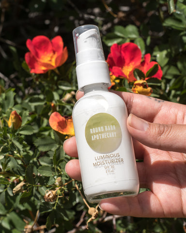 Round Barn Apothecary Luminous Moisturizer being held in a caucasian hand in front of a green leafy bush.