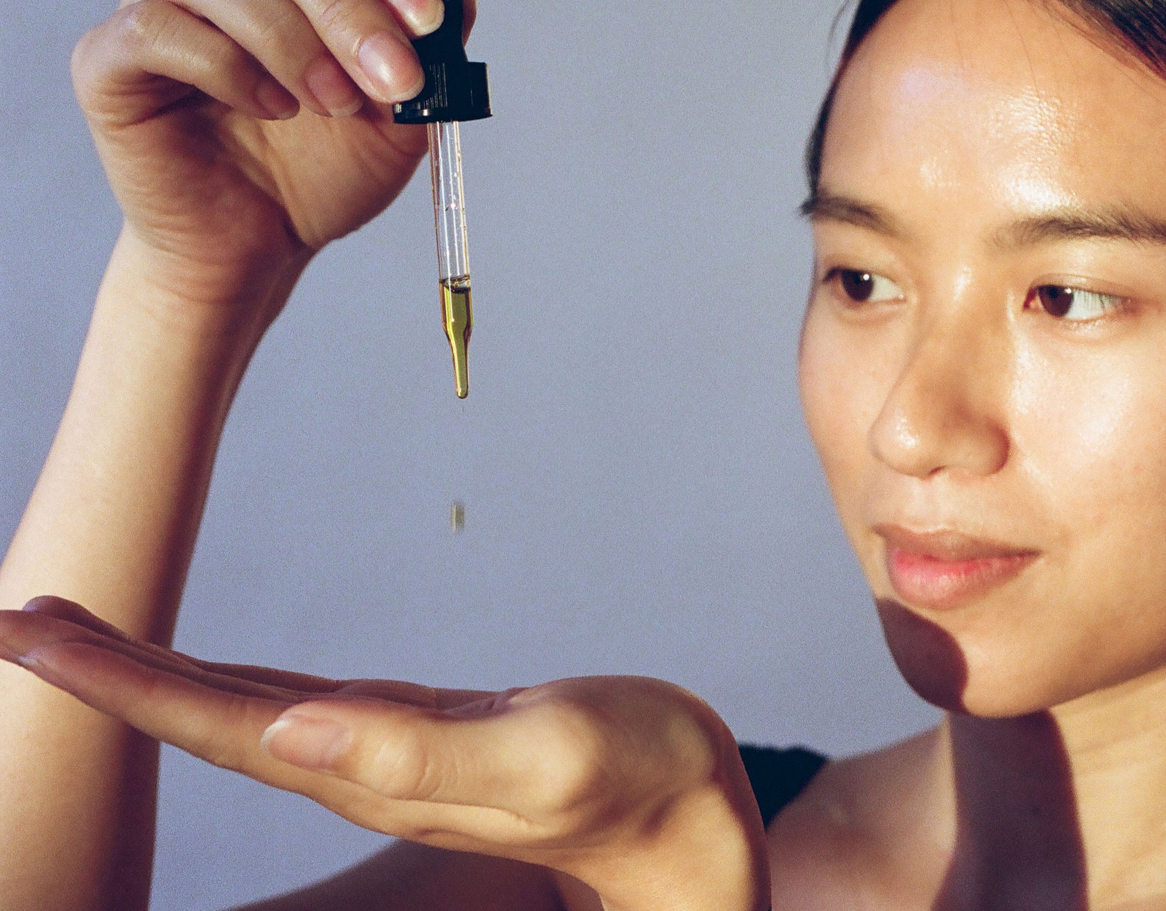 A close crop of an Asian femme person dripping face oil from a dropper into an outstretched hand