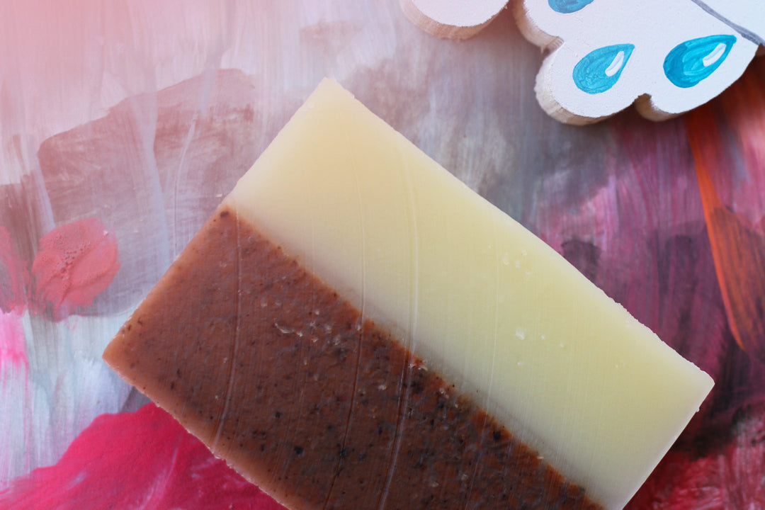 A two-tone pink and cream bar of soap sits on a pink and reddish background. There is an enlarged wooden emoji also on the table. A cloud with raindrops.