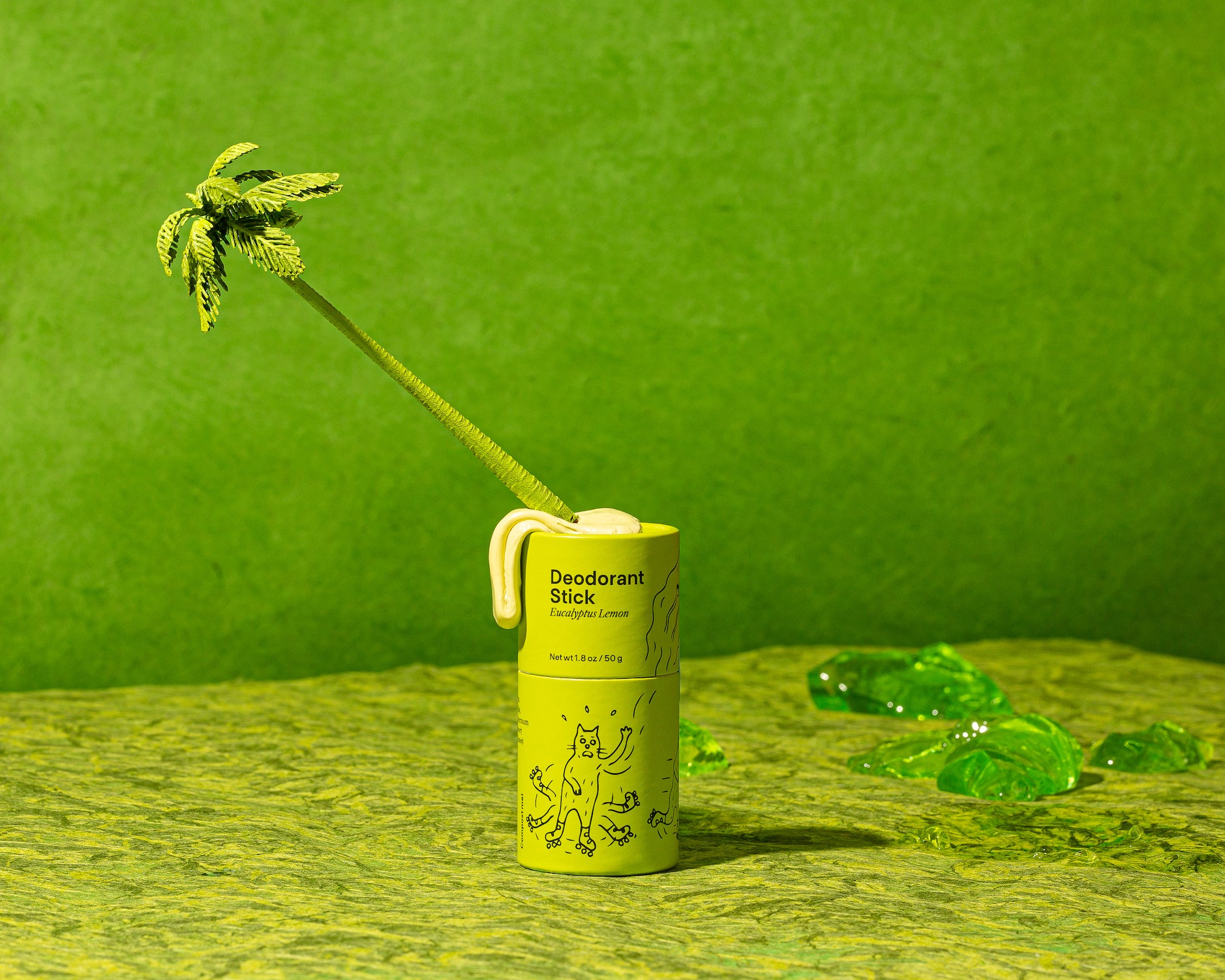 A green tube of deodorant sits on a green backdrop. A plastic palm tree is stuck diagonally out of the top.