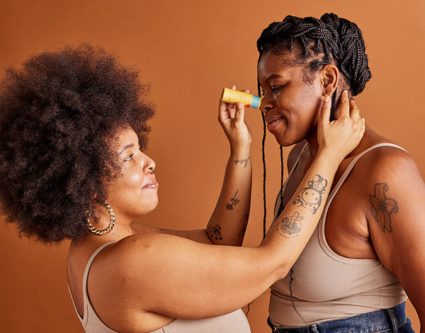 A Black woman with an afro lovingly applies balm to the nose of a taller Black woman with braids