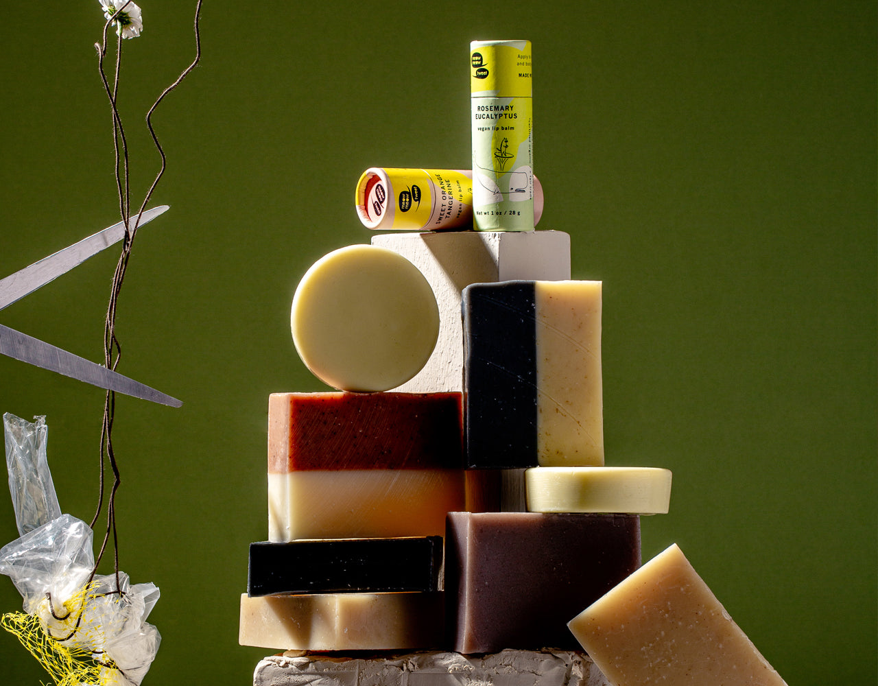 our soaps and lip balms stand in a tower against a pine backdrop, with a pair of scissors on the left open and ready to cut the stem of a flower that's connected to plastic waste