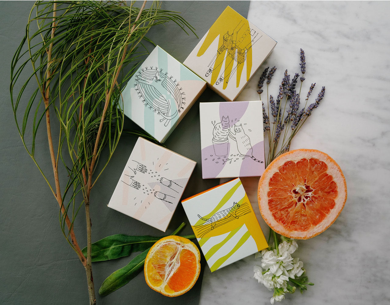 5 bars of Meow Meow Tweet soaps on a white marble surface with gray paper, surrounded by pine fronds, lavender stalks, and halved oranges