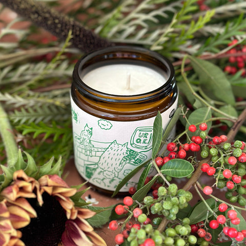 A candle in an amber jar and cream label with green drawings on it. There are plants arranged around it like holly, sunflowers and other greenery.