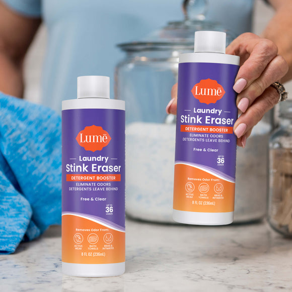 Two bottles of Laundry Stink Eraser Detergent Booster, with a woman's hand picking up one of them. There is a jar of laundry detergent in the background.