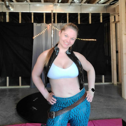 A female caucasian posing in front of a home pole. She is wearing a black blazer, white bra top, and Super Fly Honey sticky fishnet leggings.