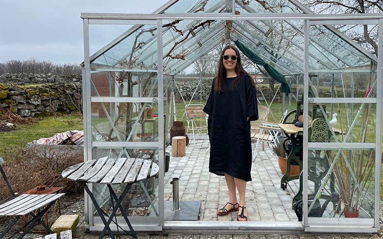 A woman stands in a greenhouse wearing a black linen kaftan