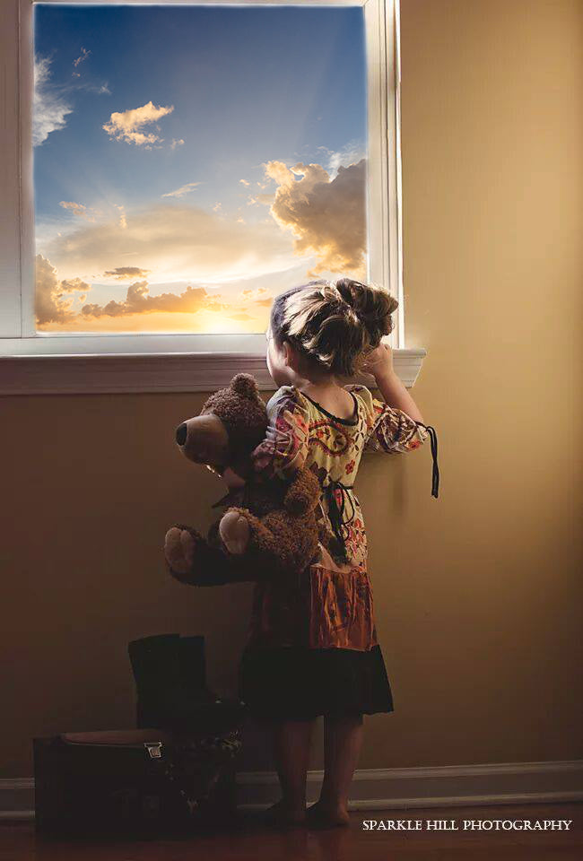 Young girl holding a teddy bear, looking out window at beautiful sunset