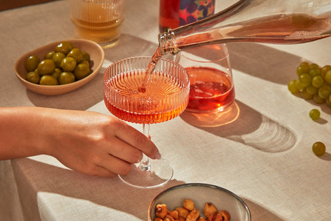 Rose Sparkler being poured into a coupe glass on a table with olives and nuts.