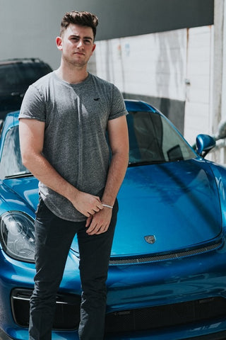 Barbat in tricou gri stand cu mainile incrucisate in fata unei masini albastre