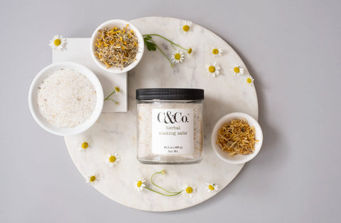 Glass jar of herbal soaking salts with three white bowls filled with raw plant material. One full of epsom salts, the other with yellow flowers petals. The background is a light grey and there are small chamomile flowers placed throughout the picture.
