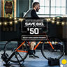 A man rides a bike on a Saris stationary bike trainer and the text is promoting $50 off select Saris trainers.