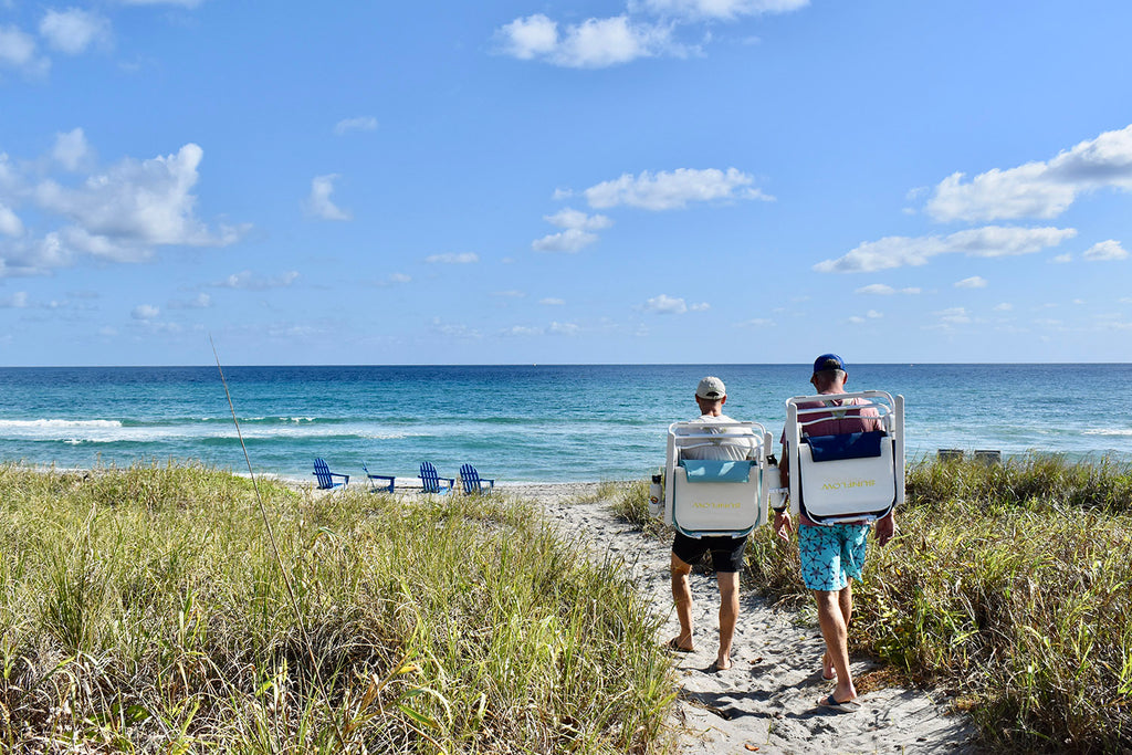 Greg and Todd walking on a sandy path toward the beach carrying SUNFLOW high tide chairs on their backs.