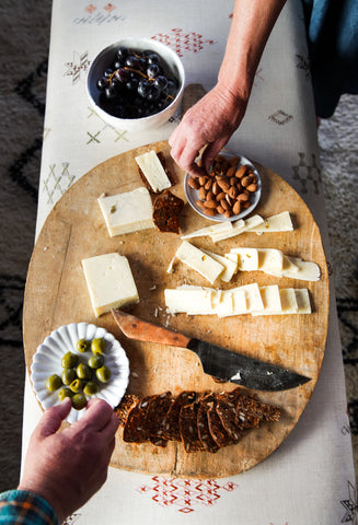 overhead view of wooden tray with various cheese, olives, and spreaders