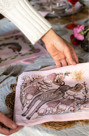 A hand at a table touching the pink Lela rose moose mushroom dinner napkin coral & Tusk collaboration