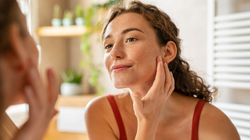 olive skintone woman with freckles in red tank top in her bathroom, looking at her reflection in the mirror and touching her face