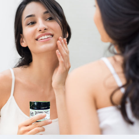 woman with long black hair looking at herself in the mirror holding a jar of erin's faces tamanu balm barrier salve to use for slugging